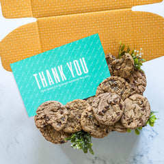 Open yellow gift box filled with assorted cookies and greenery, with a teal card that reads âThank You.â