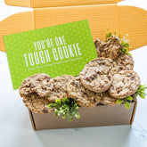 Open yellow gift box filled with assorted cookies and greenery, with a green card that reads âYouâre One Tough Cookie.â