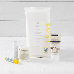 Close-up of self-care items including a âHealing Vibesâ Citrus + Sea Salt candle, goat milk hand lotion and lip balm by Beekman, refreshing face and body wipes by Beekman, and all-natural dry shampoo by Pantry, arranged on a light surface.