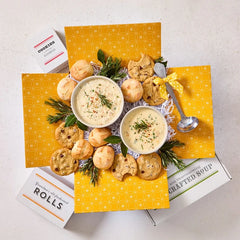 Overhead view of a yellow gift box arrangement with two bowls of creamy soup, rolls, cookies, a container labeled âHandcrafted Soup,â and a metal ladle with a yellow ribbon, arranged with greenery.
