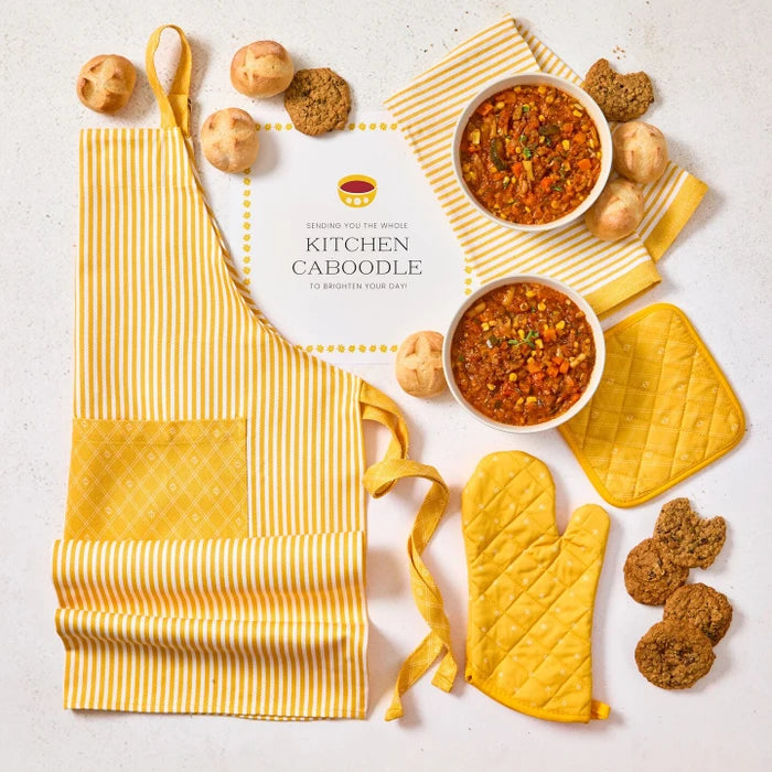 Overhead view of a kitchen-themed arrangement with two bowls of soup, rolls, cookies, a striped apron, matching oven mitts, and a card that reads âKitchen Caboodle.â