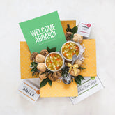 Overhead view of a yellow gift box arrangement with two bowls of soup, rolls, cookies, a container labeled âHandcrafted Soup,â a metal ladle, greenery, and a green card that reads âWelcome Aboard!â