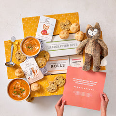 Overhead view of hands holding a card with a heartfelt message next to a comforting gift arrangement with two bowls of soup, rolls, cookies, a box labeled âHandcrafted Soup,â a box of rolls, a plush stuffed animal, childrenâs books, and a metal ladle with a yellow ribbon.