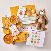 Overhead view of a yellow gift box arrangement with two bowls of soup, rolls, cookies, a container labeled âHandcrafted Soup,â a metal ladle, a stuffed animal toy, childrenâs books, and a milestone card set.