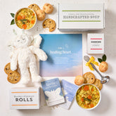 Overhead view of a sympathy-themed gift arrangement with two bowls of soup, rolls, cookies, a box labeled âHandcrafted Soup,â a metal ladle with a yellow ribbon, a plush stuffed animal, two childrenâs grief books, and a card that reads âFor a Healing Heart.â
