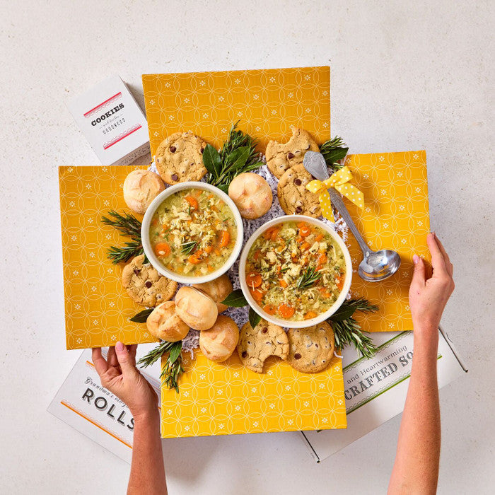 Overhead view of hands holding an open yellow gift box filled with two bowls of soup, rolls, cookies, greenery, and a metal ladle.