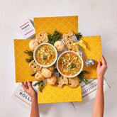 Overhead view of hands holding an open yellow gift box filled with two bowls of soup, rolls, cookies, greenery, and a metal ladle.