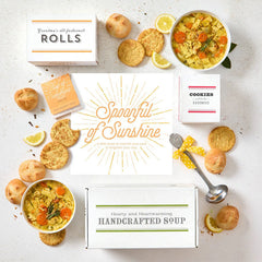 Overhead view of a sunshine-themed gift arrangement with two bowls of soup, rolls, cookies, lemon slices, a box labeled âHandcrafted Soup,â a box of rolls, a box of cookies, a metal ladle with a yellow ribbon, and a card that reads âSpoonful of Sunshine.â