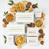 Overhead view of a symmetrical arrangement of soup gift items including four bowls of soup, rolls, cookies, multiple boxes labeled âHandcrafted Soupâ and âRolls,â and boxes of cookies arranged with greenery.