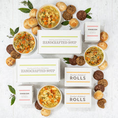 Overhead view of a symmetrical arrangement of soup gift items including four bowls of soup, rolls, cookies, multiple boxes labeled âHandcrafted Soupâ and âRolls,â and boxes of cookies arranged with greenery.