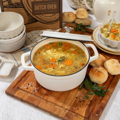 Close-up of a white Dutch oven filled with soup on a wooden board, surrounded by rolls, stacked bowls, and kitchen items in the background.