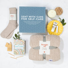 Overhead view of a self-care gift arrangement with a cozy blanket labeled âSnuggle Up,â a pair of socks, a candle, a coloring book with pencils, and a blue card that reads âSent With Care for Self-Care.â