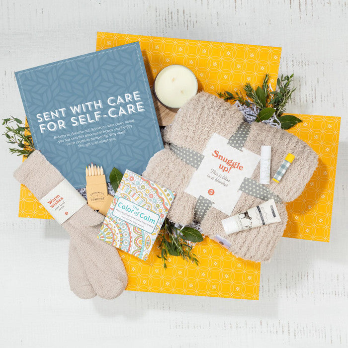 Overhead view of a yellow gift box containing a blue card that reads âSent With Care for Self-Care,â a candle, one pair of socks, small self-care items, and greenery.
