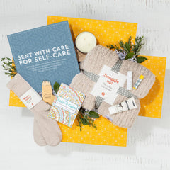 Overhead view of a yellow gift box containing a blue card that reads âSent With Care for Self-Care,â a candle, one pair of socks, small self-care items, and greenery.