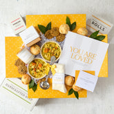 Overhead view of a yellow gift box arrangement with two bowls of soup, rolls, cookies, a container labeled âHandcrafted Soup,â a metal ladle, a candle, a pair of cozy socks, and a card that reads âYou Are Loved.â