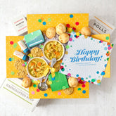 Overhead view of a birthday-themed gift arrangement with two bowls of soup, rolls, cookies, a container labeled âHandcrafted Soup,â a metal ladle with a yellow ribbon, green birthday napkins, confetti poppers, colorful confetti, and a card that reads âHappy Birthday!â