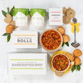 Overhead view of a soup gift arrangement with two bowls of soup, rolls, cookies, two containers of soup, a box labeled âHandcrafted Soup,â and a metal ladle with a yellow ribbon.