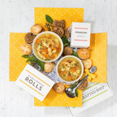 Top-down view of a get well soon gift package with two bowls of soup, dinner rolls, cookies, and a spoon arranged on a bright yellow box