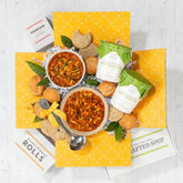 Overhead view of a yellow gift box filled with two bowls of soup, packaged rolls, two containers of soup, cookies, and a metal ladle arranged with greenery.