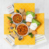 Overhead view of a yellow gift box arrangement with two bowls of soup, rolls, cookies, two containers of soup, greenery, and a metal ladle with a yellow ribbon.
