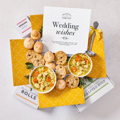 Overhead view of a yellow gift box arrangement with two bowls of soup, rolls, cookies, a container labeled âHandcrafted Soup,â a metal ladle with a yellow ribbon, and a card that reads âWedding Wishes.â