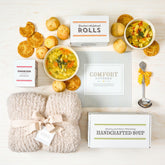 Overhead view of a comfort-themed gift arrangement with two bowls of soup, rolls, cookies, a box labeled âHandcrafted Soup,â a box of rolls, a box of cookies, a metal ladle with a yellow ribbon, a cozy knit blanket, and a card that reads âComfort Covered.â