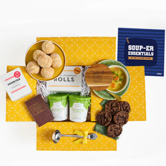 Overhead view of a yellow gift box arrangement with a bowl of soup, rolls, cookies, two containers of soup, a metal ladle, a brown book labeled âCollege Essentials,â and a blue card that reads âSoup-er Essentials.â