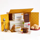 Open yellow gift box containing packaged rolls, a container labeled âHandcrafted Soup,â a box of cookies, a stack of cookies, rolls on a plate, a metal ladle with a yellow ribbon, and a small book.
