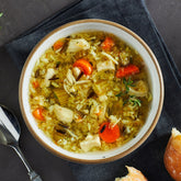 Close-up of a bowl of chicken and wild rice soup with carrots, herbs, and shredded chicken, placed beside a spoon and a piece of bread.