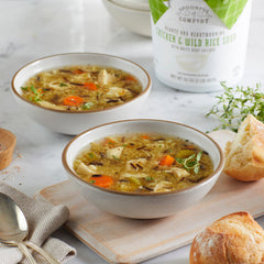 Close-up of two bowls of chicken and wild rice soup with carrots and herbs, served with pieces of bread on a wooden board, with a soup container in the background.