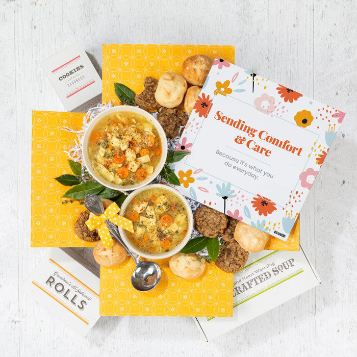 Overhead view of a yellow gift box arrangement with two bowls of soup, rolls, cookies, a container labeled âHandcrafted Soup,â a metal ladle with a yellow ribbon, and a floral card that reads âSending Comfort & Care.â