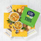 Overhead view of a yellow gift box arrangement with two bowls of soup, rolls, cookies, a container labeled âHandcrafted Soup,â a metal ladle with a yellow ribbon, and a green card that reads âYouâve Got This.â