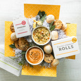 Overhead view of a yellow gift box arrangement with two bowls of soup, rolls, cookies, a container labeled âHandcrafted Soup,â and packaged rolls, arranged with greenery.