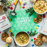 Overhead view of a birthday-themed food gift arrangement with bowls of soup, cookies, rolls, and packaged cookies surrounding a green card that reads âBirthday Dinner Our Treat,â with colorful confetti scattered on the table.