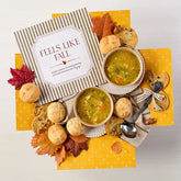 Overhead view of a fall-themed arrangement with two bowls of soup, rolls, cookies, a box labeled âHandcrafted Soup,â a box of rolls, a metal ladle with a yellow ribbon, autumn leaves, and a card that reads âFeels Like Fall.â