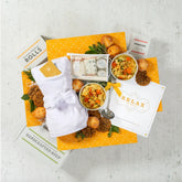 Overhead view of a relaxation-themed gift arrangement with two bowls of soup, rolls, cookies, a box labeled âHandcrafted Soup,â a robe, self-care items in a toiletry bag, a metal ladle with a yellow ribbon, and a card that reads âRelax.â