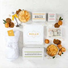 Overhead view of a relaxation-themed gift arrangement with two bowls of soup, rolls, cookies, a box labeled âHandcrafted Soup,â a robe, self-care items in a toiletry bag, a metal ladle, and a card that reads âRelax.â