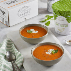 Close-up of two bowls of tomato basil soup garnished with cream and basil, with a striped towel, spoon, and a soup container with a green lid in the background.