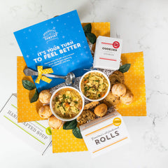 Overhead view of a yellow gift box arrangement with two bowls of soup, rolls, cookies, a container labeled âHandcrafted Soup,â a metal ladle with a yellow ribbon, and a blue card that reads âItâs Your Turn to Feel Better.â