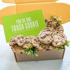 Open yellow gift box filled with assorted cookies and greenery, with a green card that reads âYouâre One Tough Cookie.â