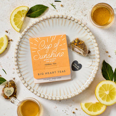 Close-up of a package of âCup of Sunshineâ herbal tea placed on a plate, surrounded by lemon slices, tea, a honey dipper, and small decorative items.