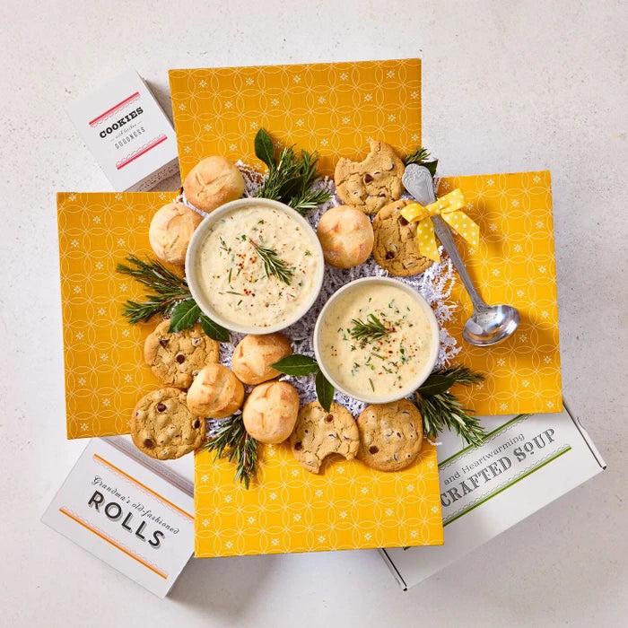 Overhead view of a yellow gift box arrangement with two bowls of creamy soup, rolls, cookies, a container labeled âHandcrafted Soup,â and a metal ladle with a yellow ribbon, arranged with greenery.