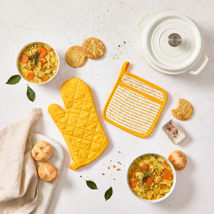 Overhead view of a kitchen-themed arrangement with two bowls of soup, rolls, cookies, a yellow oven mitt and pot holder, a white Dutch oven, and a towel on a light surface.