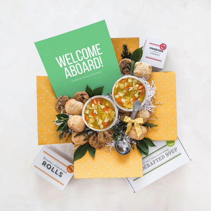 Overhead view of a yellow gift box arrangement with two bowls of soup, rolls, cookies, a container labeled âHandcrafted Soup,â a metal ladle, greenery, and a green card that reads âWelcome Aboard!â