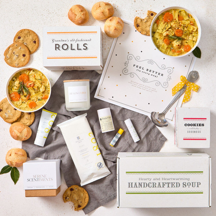 Overhead view of a get-well-themed gift arrangement with two bowls of soup, rolls, cookies, a box labeled âHandcrafted Soup,â a box of rolls, self-care items including lotion, lip balm, wipes, and dry shampoo, a candle, a metal ladle with a yellow ribbon, and a card with a get-well message.