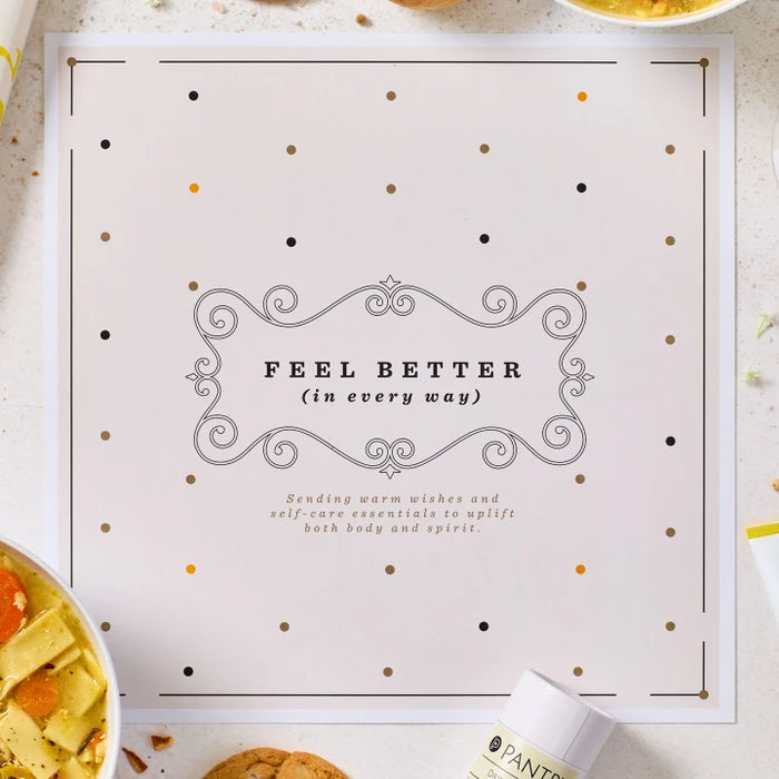 Close-up of a card that reads âFeel Better (in every way),â with decorative accents and parts of a soup and self-care gift arrangement visible around it.
