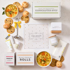 Overhead view of a get-well-themed gift arrangement with a bowl of soup, rolls, cookies, a box labeled âHandcrafted Soup,â a box of rolls, self-care items including a candle and lotion, a metal ladle with a yellow ribbon, and a card with a get-well message.