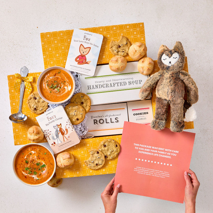 Overhead view of hands holding a card with a heartfelt message next to a comforting gift arrangement with two bowls of soup, rolls, cookies, a box labeled âHandcrafted Soup,â a box of rolls, a plush stuffed animal, childrenâs books, and a metal ladle with a yellow ribbon.
