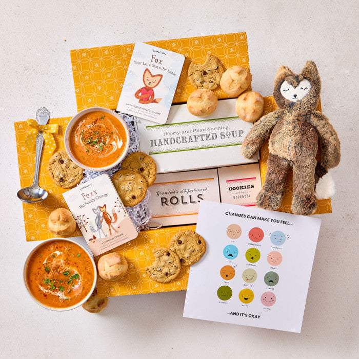 Overhead view of a yellow gift box arrangement with two bowls of soup, rolls, cookies, a container labeled âHandcrafted Soup,â a metal ladle, a stuffed animal toy, childrenâs books, and a milestone card set.