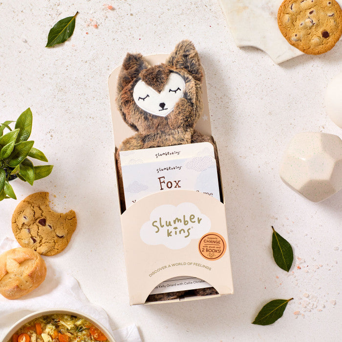Close-up of a âSlumberkinsâ plush fox toy in its box, surrounded by cookies, rolls, greenery, and a bowl of soup partially visible nearby.